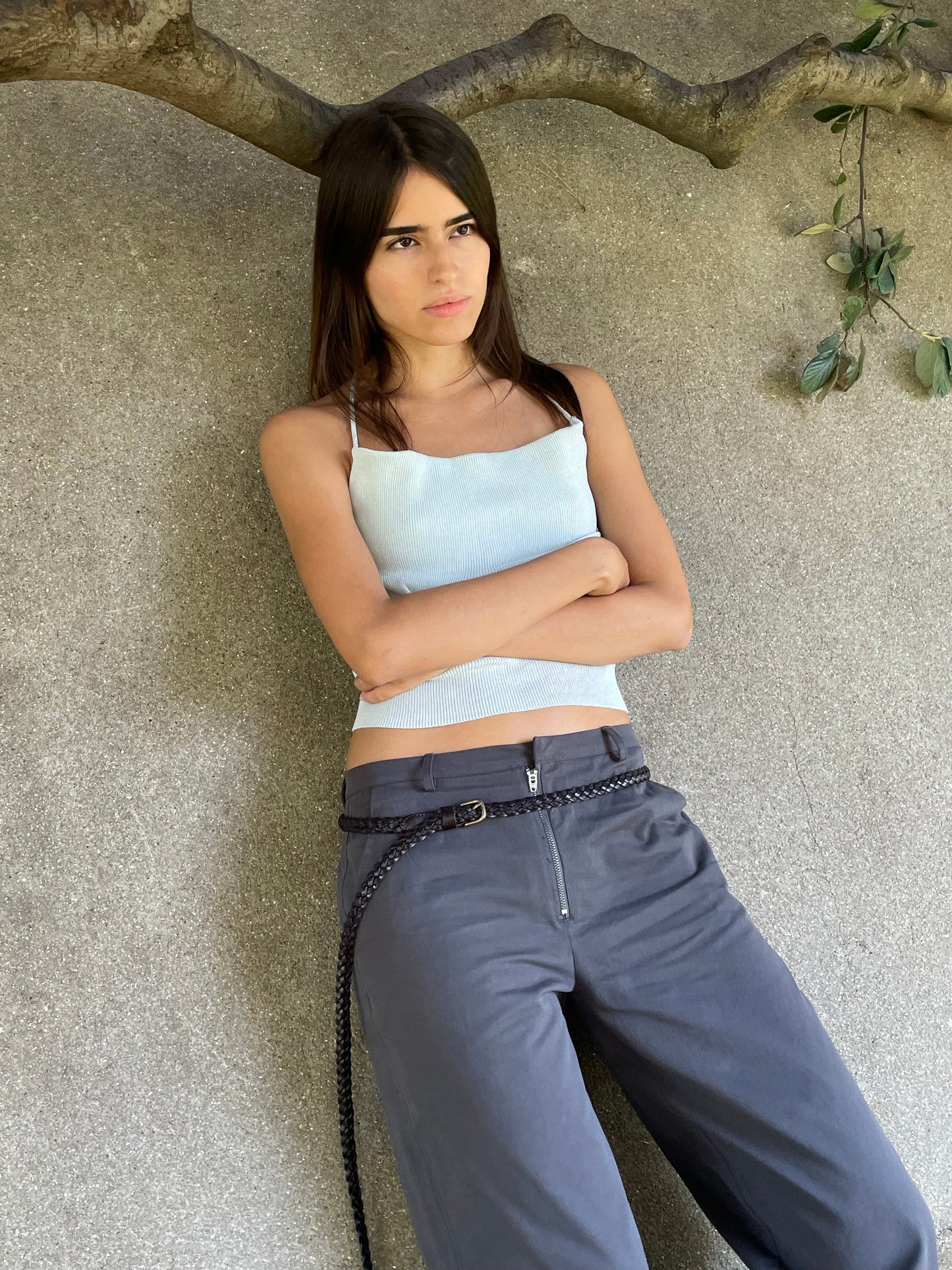 A young woman with long dark hair stands against a textured gray wall beneath a tree branch, wearing the Zip Pant - Charcoal by Deiji Studios with a white sleeveless top and braided belt, arms crossed and gazing to the side.