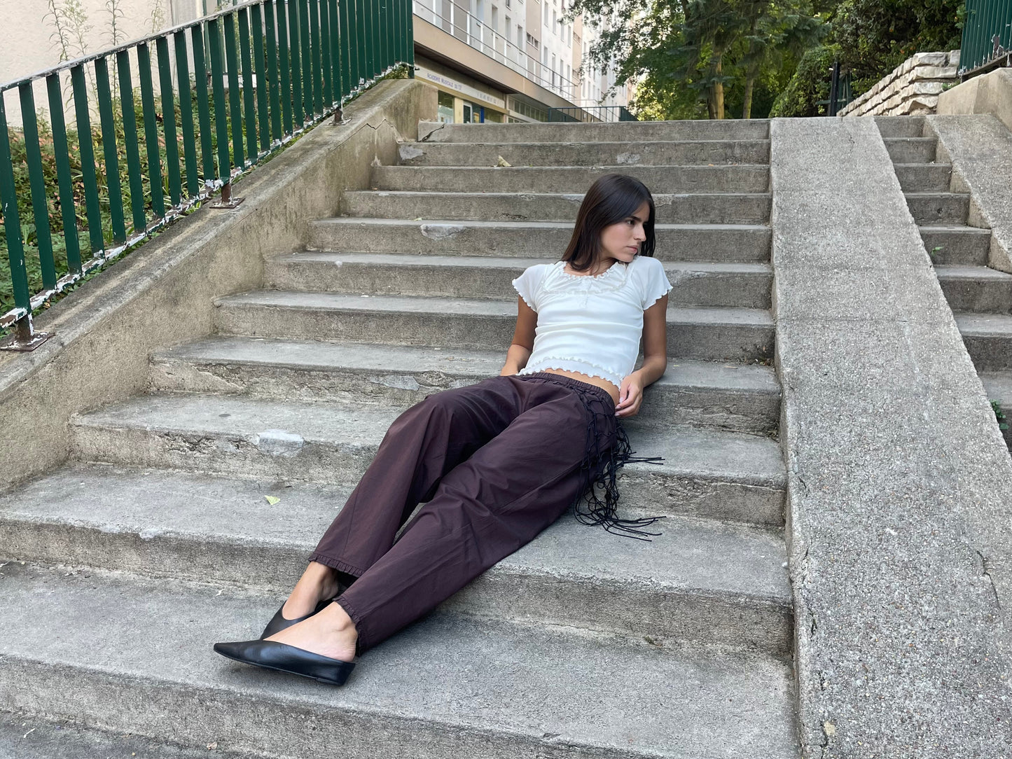 A woman with straight brown hair sits on outdoor concrete steps, wearing a white organic cotton top, black pointed shoes, and the Flounce Pant - Umber by Deiji Studios. She looks to her left, with greenery and buildings behind her.