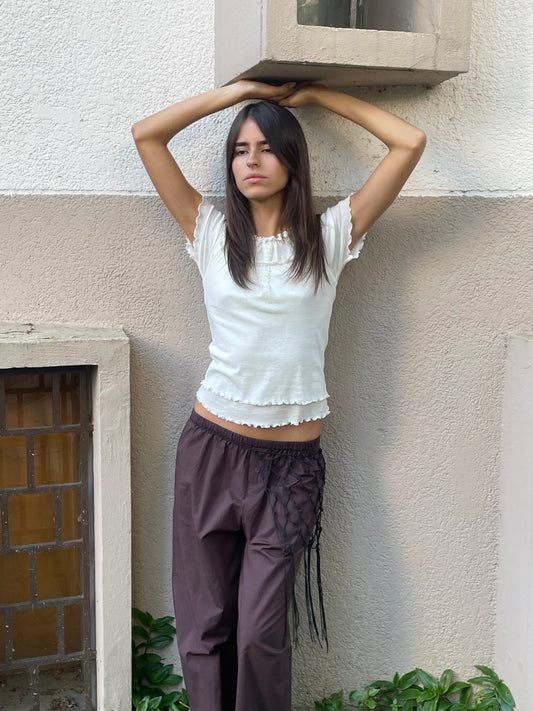 A young woman with straight dark hair stands against a beige and white wall, wearing a white ruffled top and Deiji Studios' Flounce Pant - Umber. She rests her arms above her head, looking calmly at the camera with green plants nearby.
