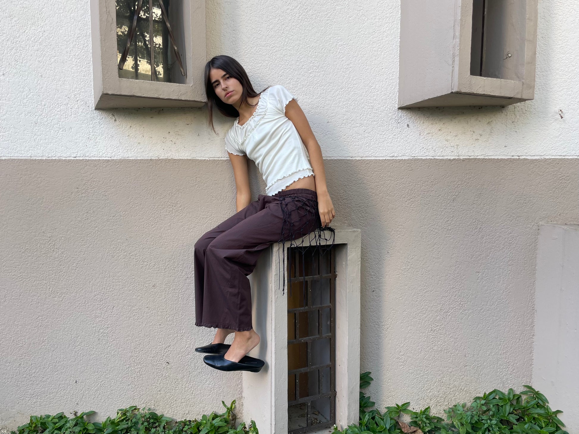 A young woman with straight dark hair sits on a small rectangular structure against a two-toned wall, wearing a white Deiji Studios top, Flounce Pant - Umber, and black shoes, with greenery at the wall's base.