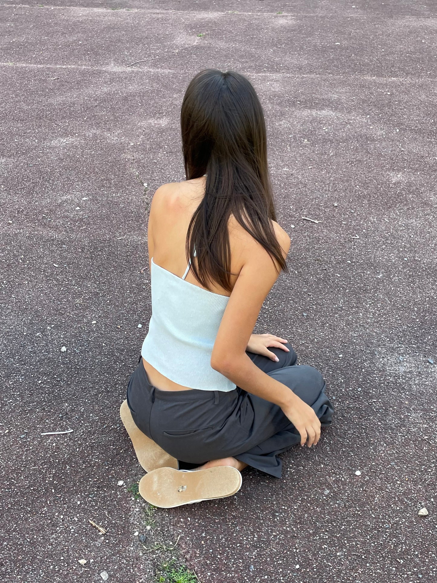 A young woman with long brown hair sits on textured dark pavement, facing away in a Fine Knot Tank - Ice Blue from Deiji Studios, paired with dark pants and beige sandals.