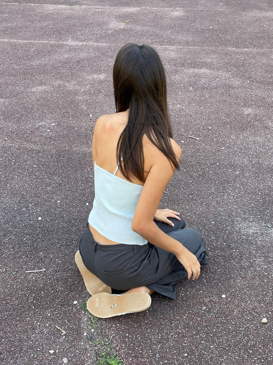 A young woman with long brown hair sits on textured dark pavement, facing away in a Fine Knot Tank - Ice Blue from Deiji Studios, paired with dark pants and beige sandals.