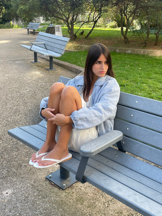 A young woman sits on a gray park bench, knees to her chest, wearing the Double Layered Jacket - Blue Stripe over a white dress with white flip-flops. She gazes thoughtfully to the side, surrounded by trees and grass.