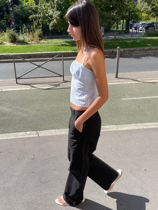 A woman with long brown hair walks on a sunny street, wearing the Strap Top - Mineral Stripe, black wide-leg pants, and white sandals. Trees and cars are visible in the background.