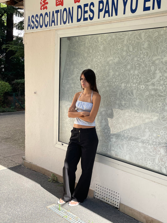 A woman with long dark hair, wearing a light blue crop top, Zip Pant - Black, and white sandals stands with arms crossed in front of a building featuring a large window and an ASSOCIATION DES PAN YU EN sign.