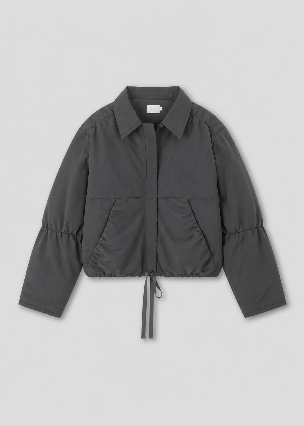 The Gathered Jacket - Slate by Deiji Studios is a charcoal gray, lightweight, oversized jacket featuring long sleeves, a collar, front zip closure, side pockets, and an adjustable drawstring hem, shown on a plain light background.