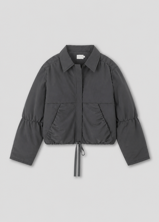 The Gathered Jacket - Slate by Deiji Studios is a charcoal gray, lightweight, oversized jacket featuring long sleeves, a collar, front zip closure, side pockets, and an adjustable drawstring hem, shown on a plain light background.