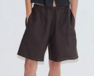 A person wearing the Half Short in a loose-fitting, dark brown style with hands in pockets, standing against a plain light background. The upper body is partially visible and features a black top.