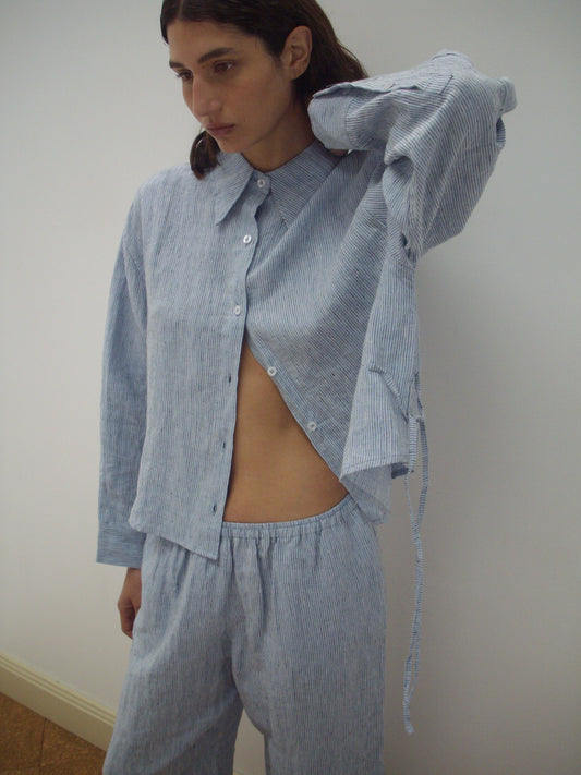 A person with shoulder-length dark hair wears Deiji Studios' 4 Tie Set - Blue Stripe, featuring a loose, light blue and white striped organic shirt and matching pants. Part of the shirt is unbuttoned with one arm inside, standing against a plain background.