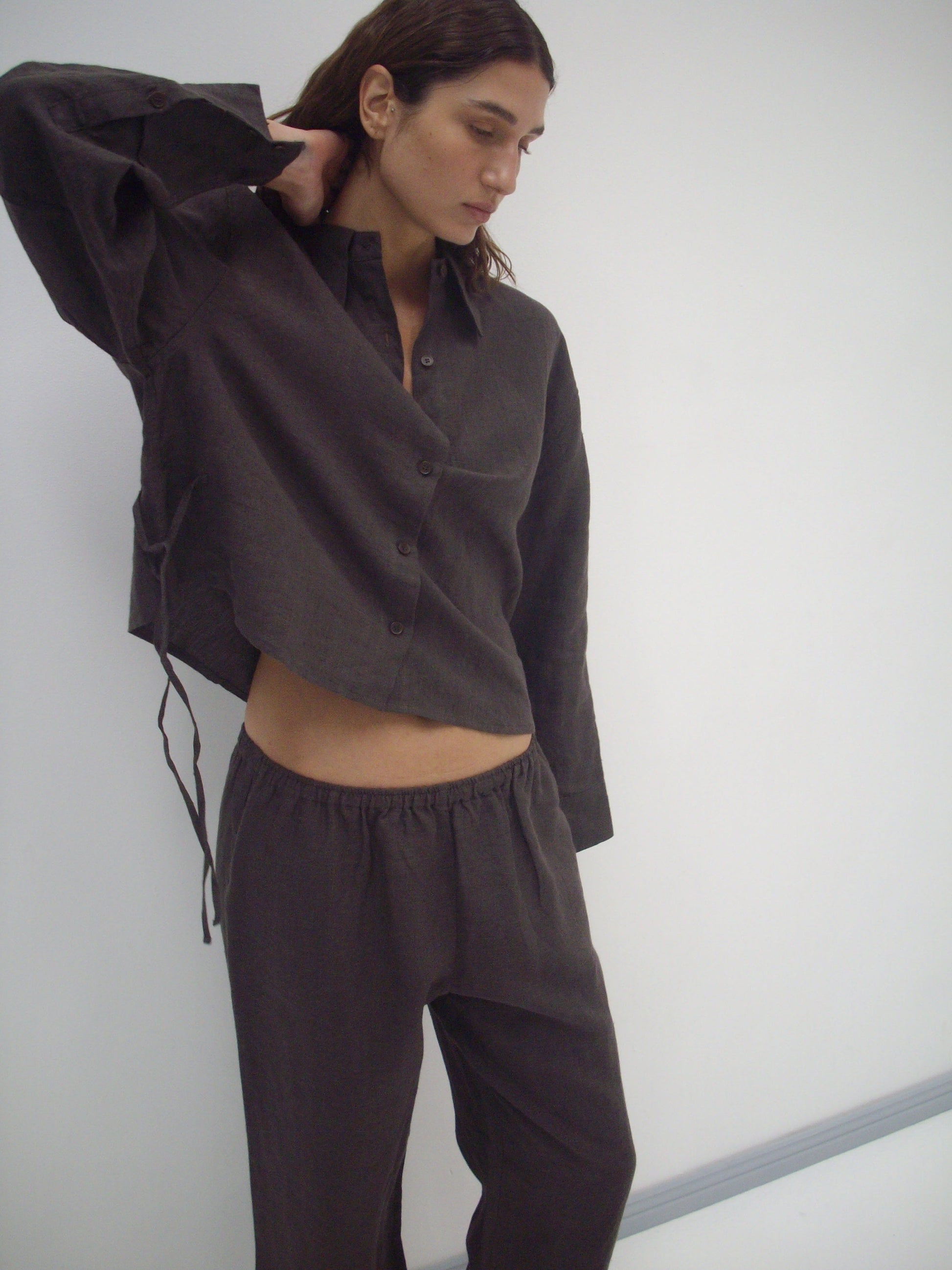 A person wears the Deiji Studios 4 Tie Set - Bark, a loose organic linen outfit in dark brown, standing against a plain white background with one arm raised and head slightly tilted down.
