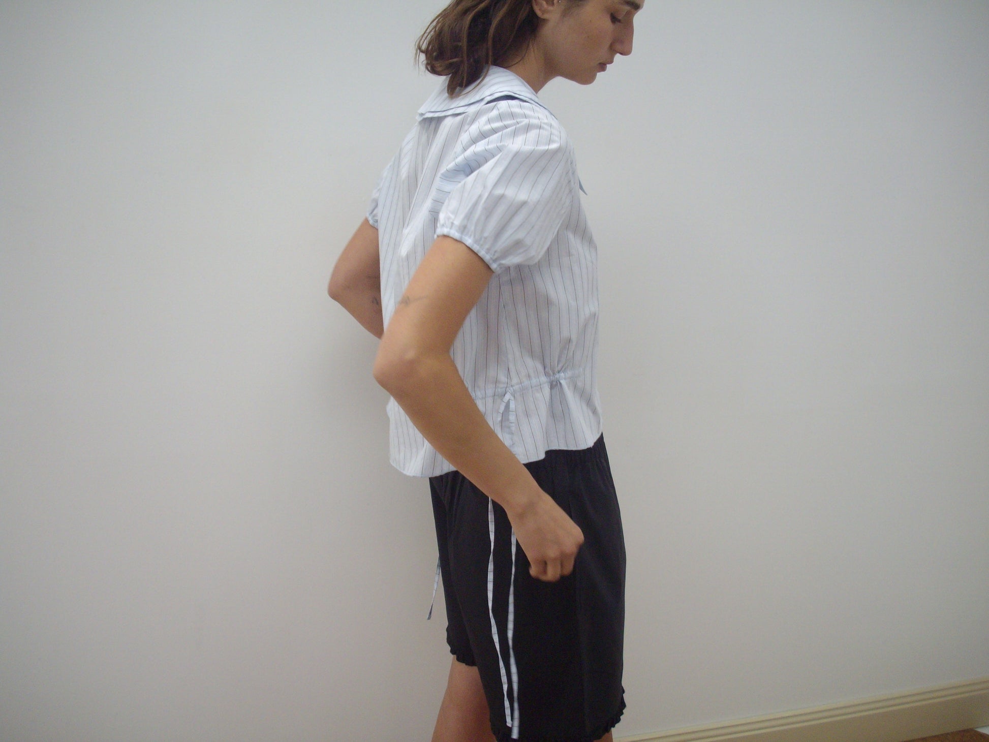 A person stands sideways against a plain white wall, wearing the Deiji Studios Double Collar Shirt - Mineral Stripe and black shorts with white side stripes.
