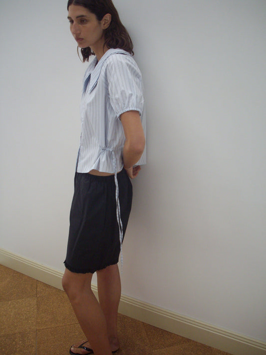 A woman with shoulder-length brown hair stands sideways against a white wall, wearing a Deiji Studios Double Collar Shirt - Mineral Stripe with short sleeves, a dark knee-length skirt, and sandals.