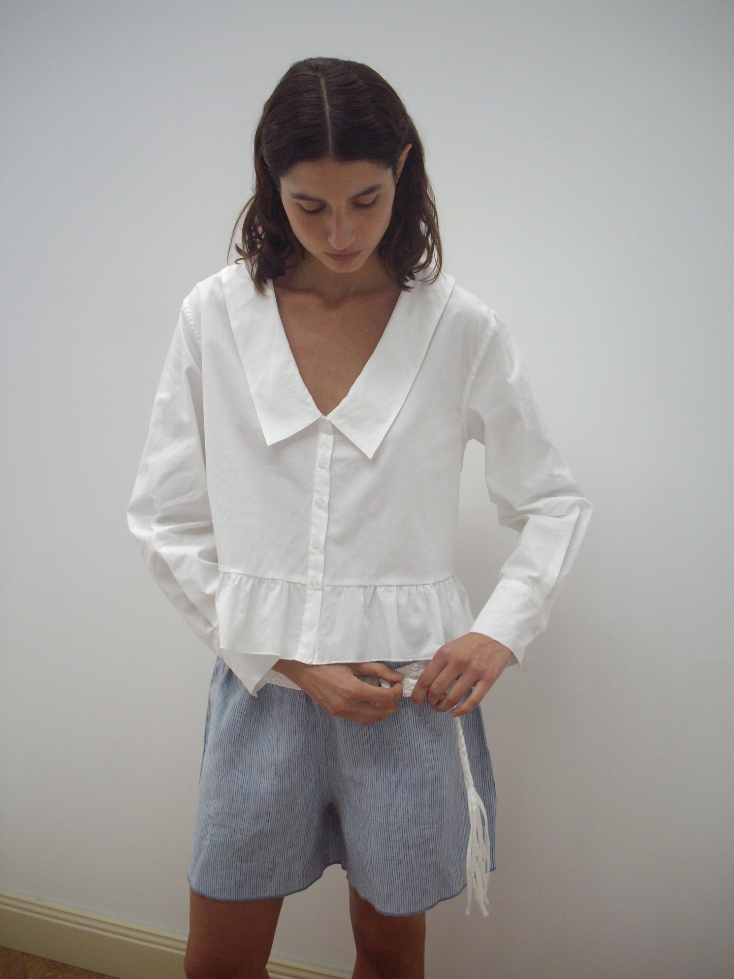 A person with shoulder-length dark hair wears the Sweep Shirt - White from Deiji Studios and light blue striped shorts, standing against a plain white wall and looking down while adjusting the shorts.