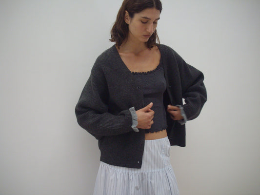 A woman stands against a white background, wearing the Flounce Cardigan - Charcoal over a matching gray top and a light pleated skirt. She looks down, adjusting her cardigan with both hands.