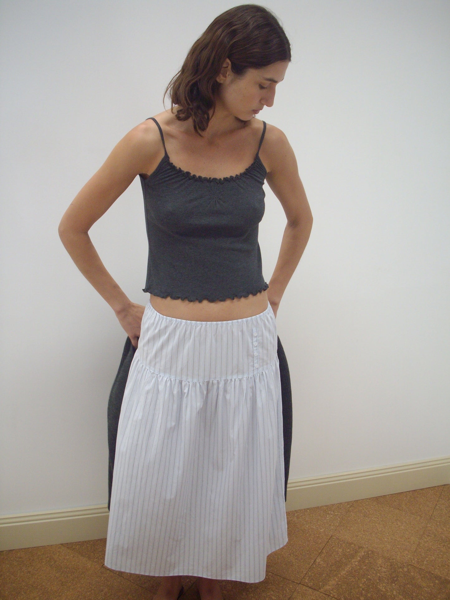 A person wears the Deiji Studios Button Skirt - Mineral Stripe, a white, knee-length skirt with thin vertical stripes, styled with a dark gray sleeveless top, standing indoors against a plain white wall and looking down to their left.