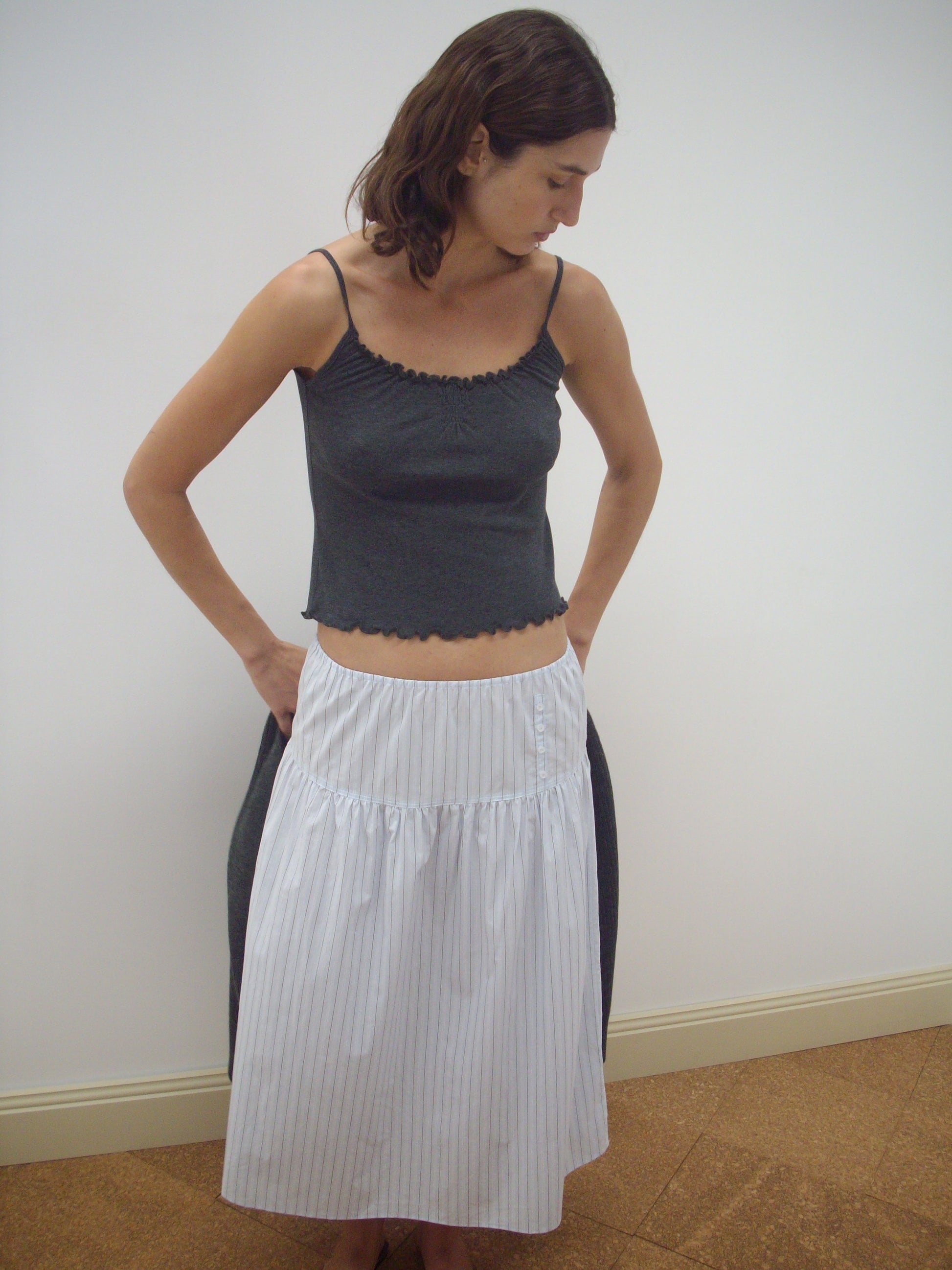 A person wears the Deiji Studios Button Skirt - Mineral Stripe, a white, knee-length skirt with thin vertical stripes, styled with a dark gray sleeveless top, standing indoors against a plain white wall and looking down to their left.