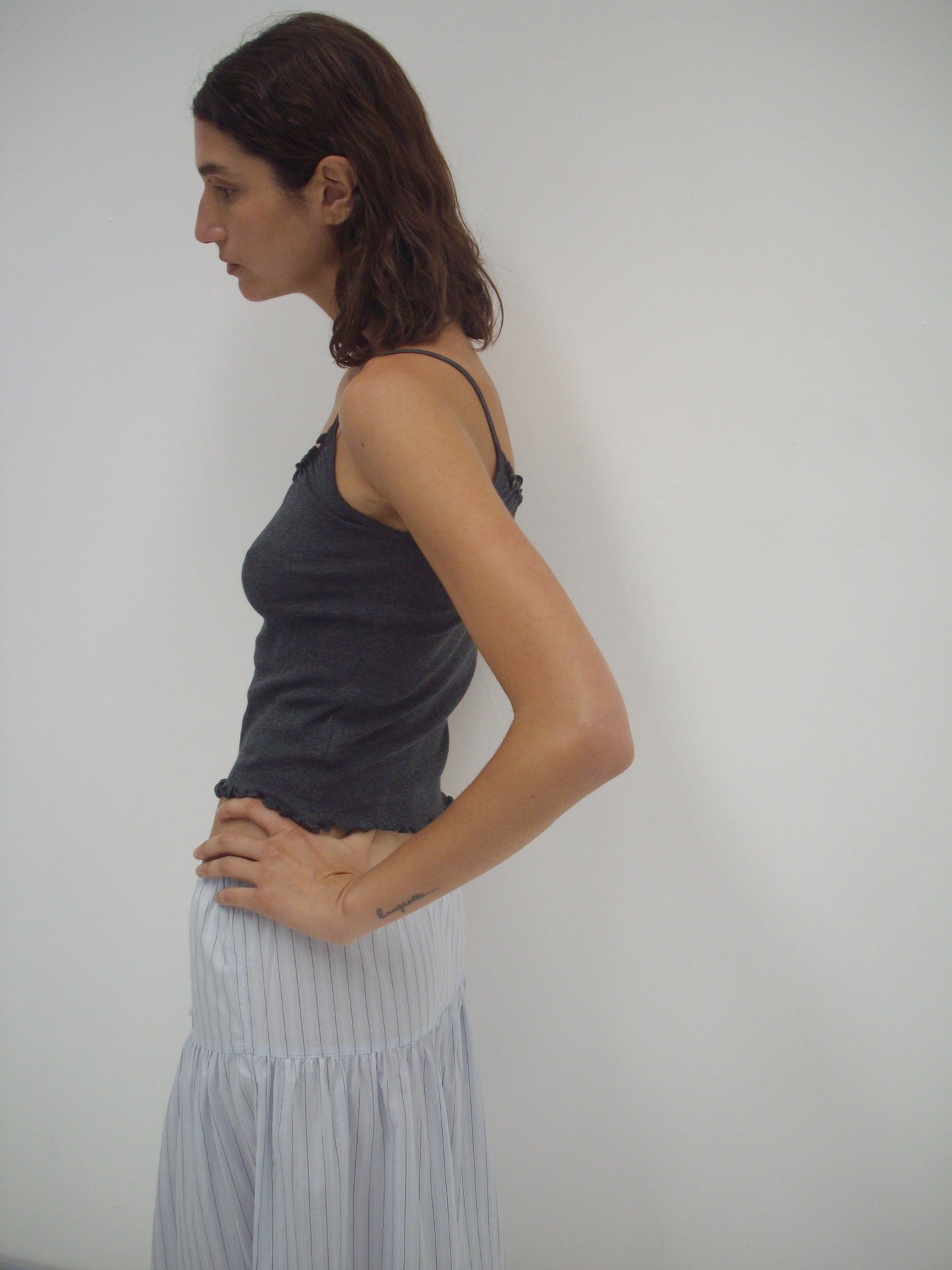 A person with shoulder-length brown hair stands in profile against a plain white background, wearing a dark sleeveless top and the Button Skirt - Mineral Stripe from Deiji Studios—a light, flowy organic cotton skirt with thin vertical stripes.