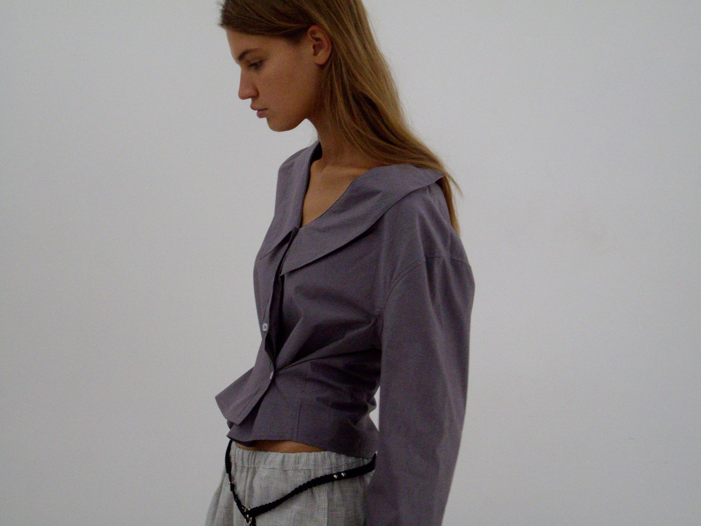 A woman with long brown hair stands in profile against a plain background, wearing the Waist Button Shirt - Currant by Deiji Studios and light gray pants, with a black beaded accessory around her waist.