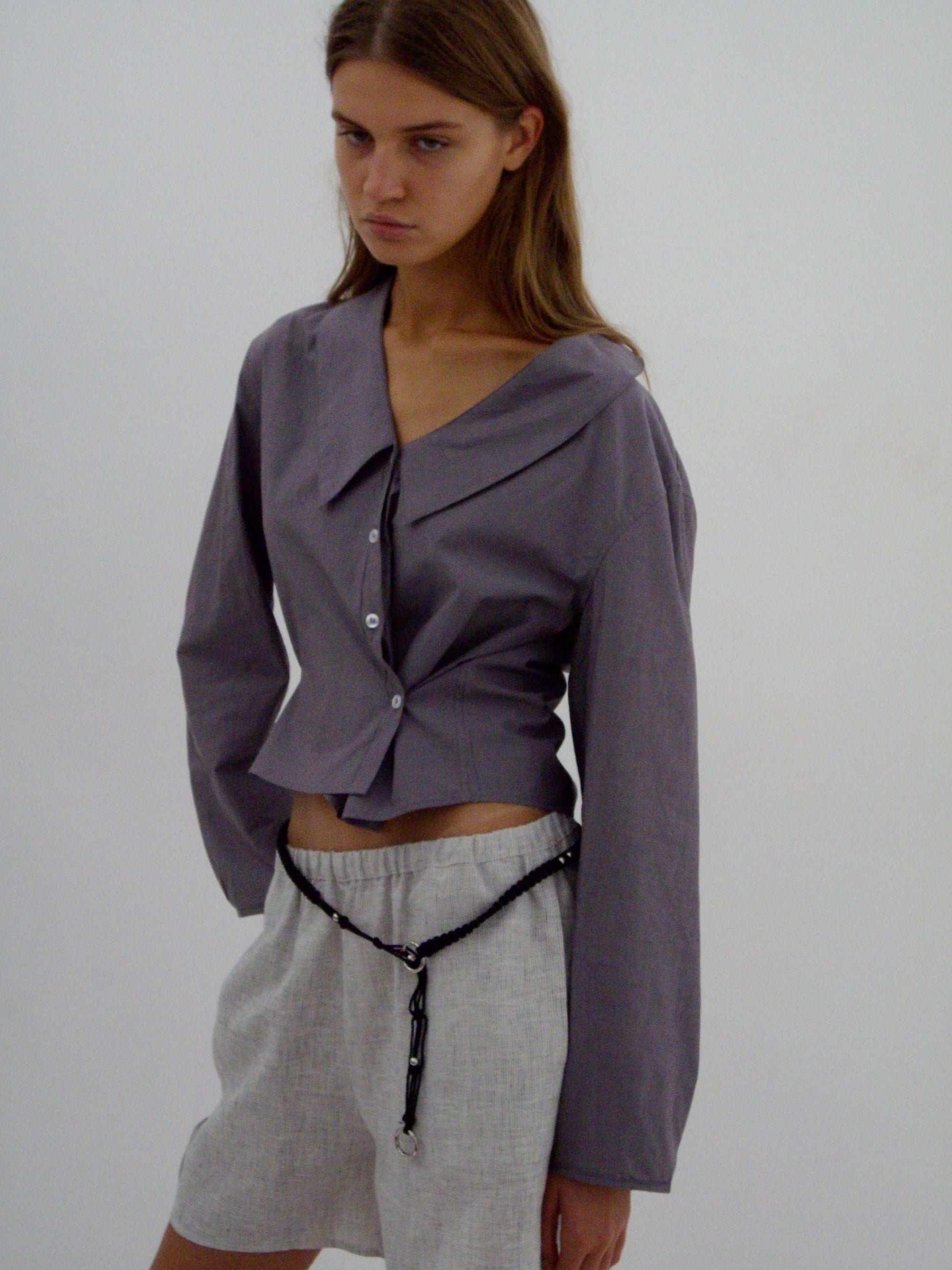A woman stands against a white background wearing a Deiji Studios Waist Button Shirt - Currant with long sleeves and light gray shorts, accessorized with a thin black belt hanging loosely around her hips.
