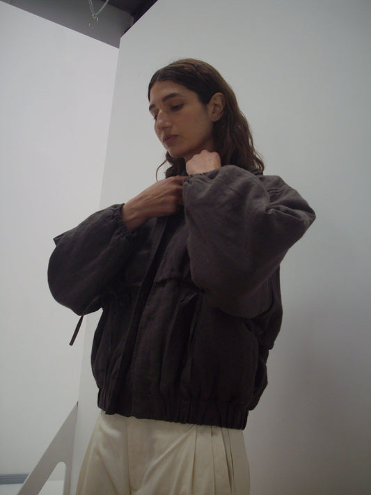 A person with long brown hair adjusts the collar of the Double Layered Jacket - Bark while standing against a plain white wall and wearing light-colored pants.