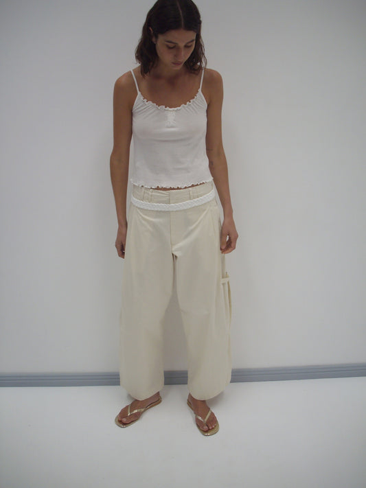 A woman stands indoors against a plain white wall, wearing Deiji Studios’ Tab Pant in off-white, paired with a white camisole and gold sandals. She looks down with her hands by her sides.