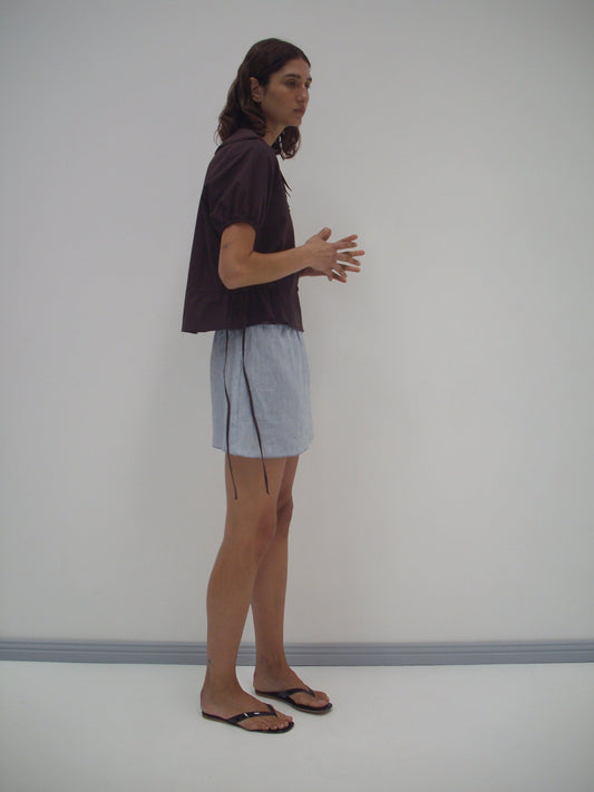 A person with long hair stands sideways against a white wall, wearing a Deiji Studios Double Collar Shirt - Umber with short sleeves, light blue shorts, and black sandals. Their hands are raised in mid-gesture.