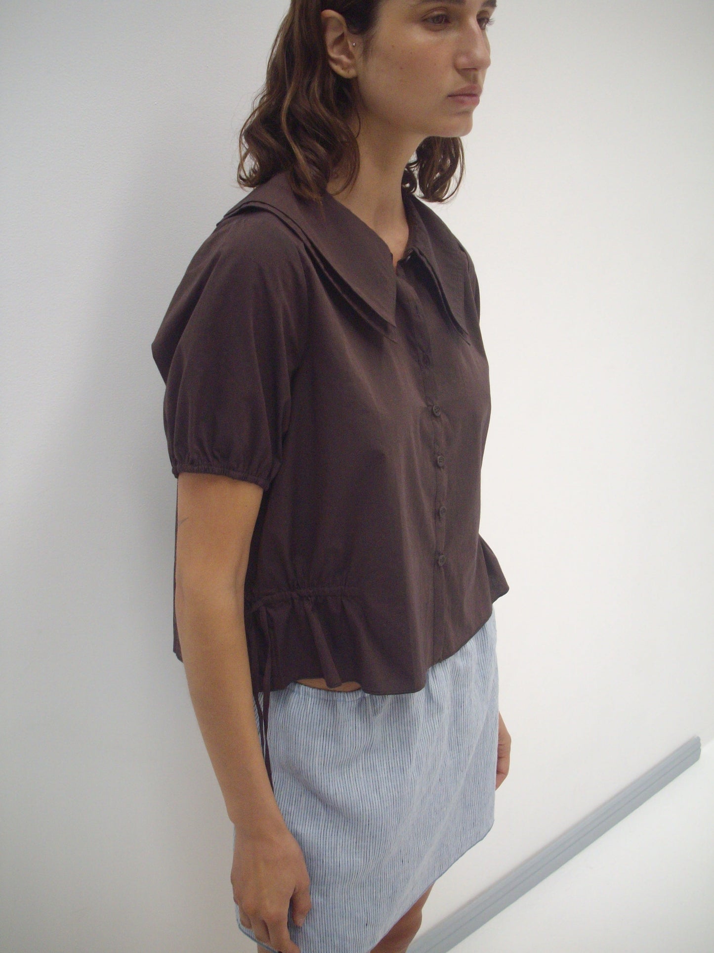 A woman with medium-length brown hair and a neutral expression stands against a plain white wall, wearing the Deiji Studios Double Collar Shirt in Umber paired with a light blue pinstriped skirt.