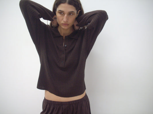 A person with wavy brown hair, wearing Deiji Studios' Polo Knit in Dark Almond and matching pants, stands against a plain white background with their hands behind their head, looking to the side.