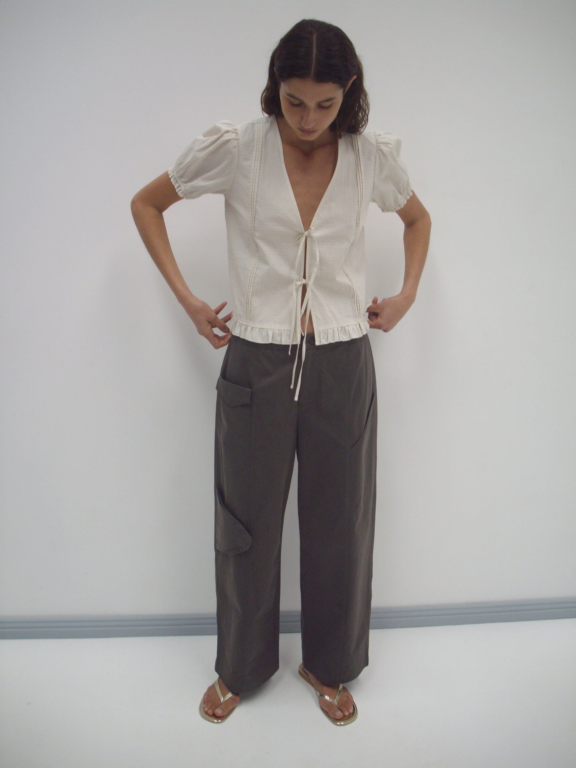 A woman stands against a plain white wall, looking down. She wears the Pintuck Top - Bone Check by Deiji Studios, featuring organic cotton with a deep V-neck and tie front, paired with loose gray pants and gold sandals.