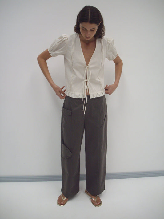 A woman stands against a plain white wall, looking down. She wears the Pintuck Top - Bone Check by Deiji Studios, featuring organic cotton with a deep V-neck and tie front, paired with loose gray pants and gold sandals.