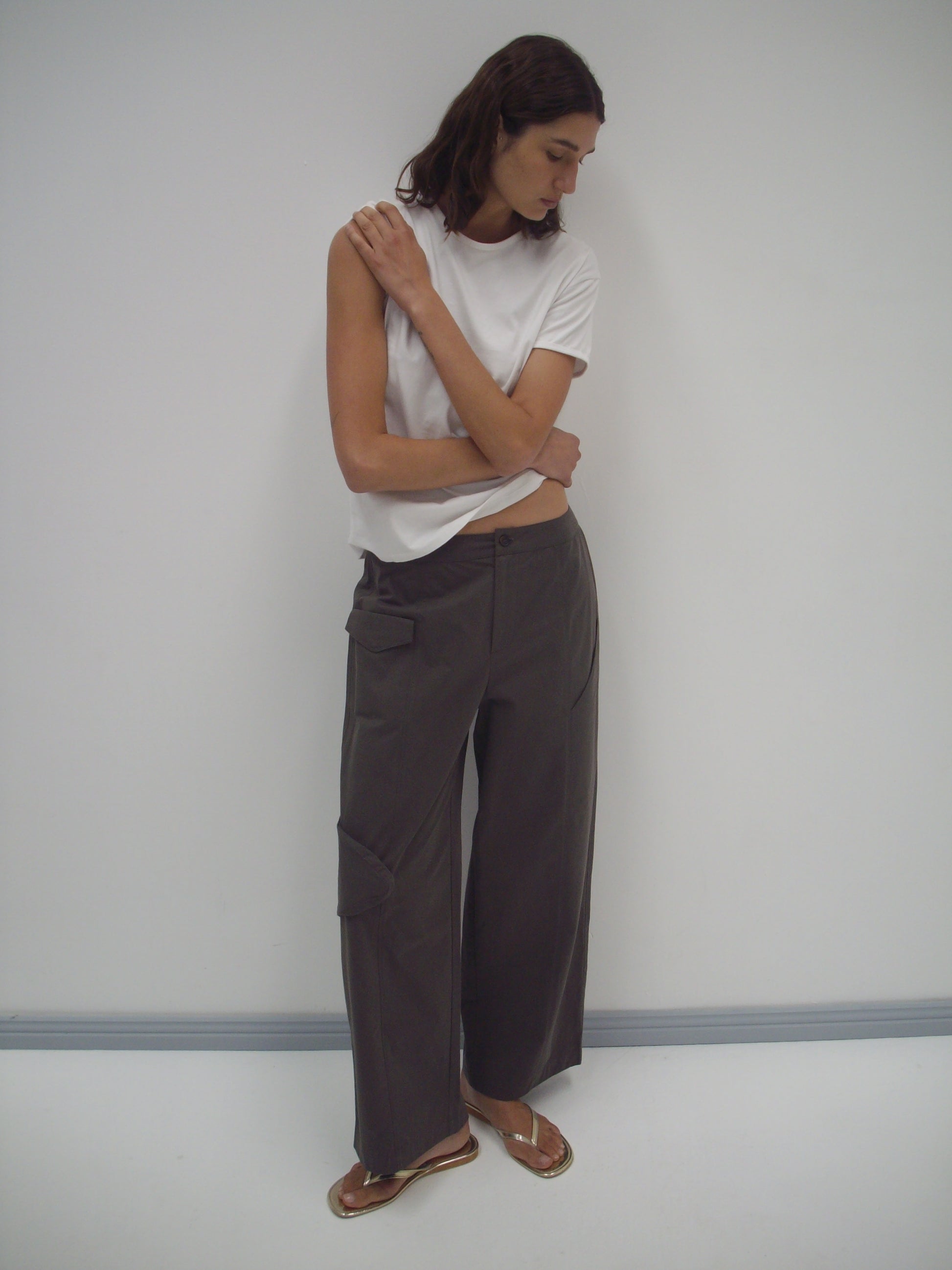A woman stands against a plain white wall, wearing the Pocket Pant - Wood in organic cotton twill. The loose, dark gray wide-leg pants feature pockets, styled with a white t-shirt and brown sandals. She looks down, one arm bent to her shoulder.