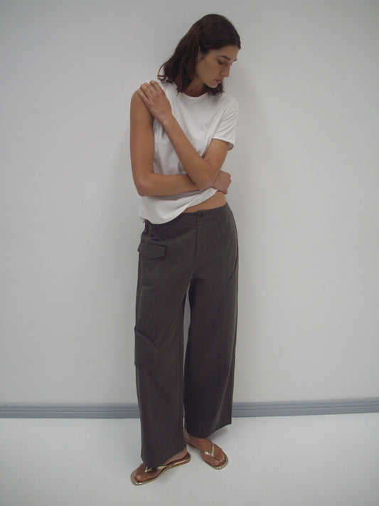 A woman stands against a plain white wall, wearing the Pocket Pant - Wood in organic cotton twill. The loose, dark gray wide-leg pants feature pockets, styled with a white t-shirt and brown sandals. She looks down, one arm bent to her shoulder.