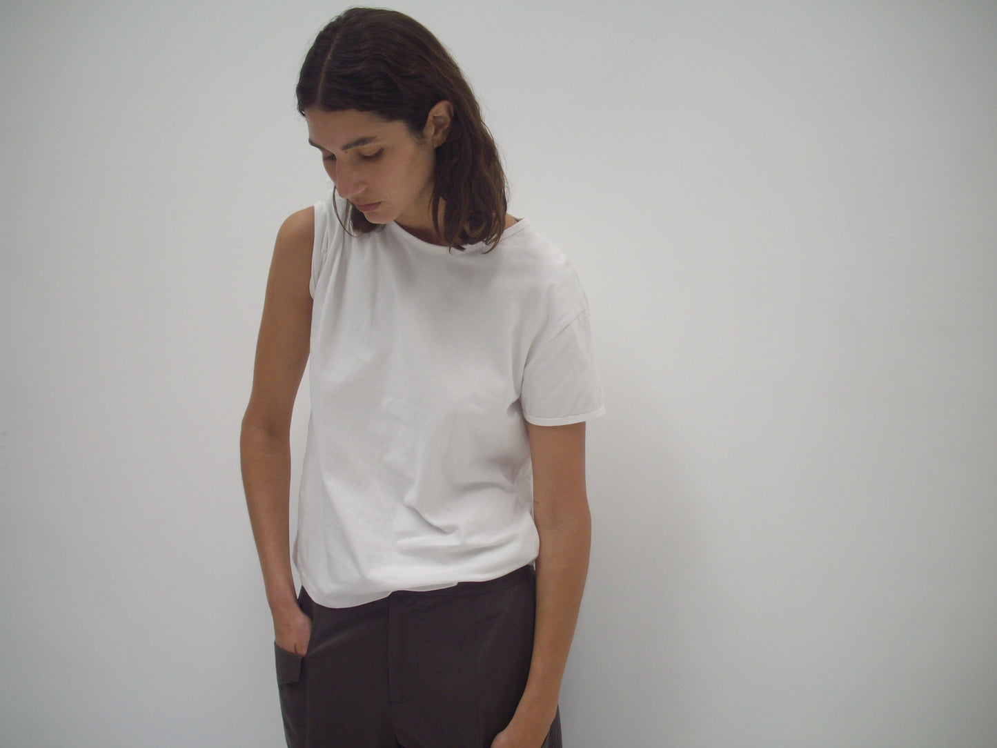 A person with shoulder-length brown hair wears the Simple Tee - White by Deiji Studios and dark pants, standing against a blank white wall and looking down with one hand in their pocket.