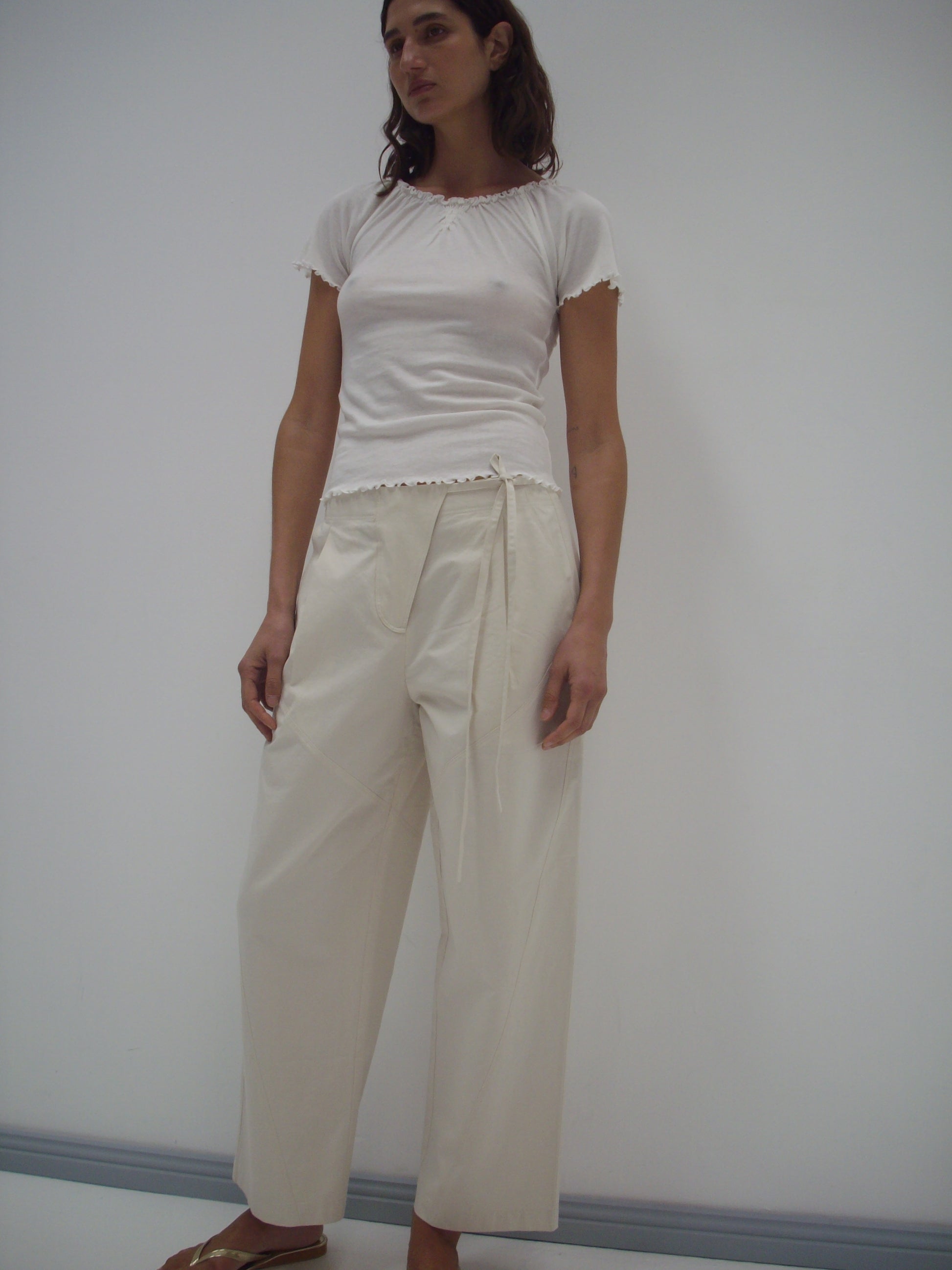 A woman with shoulder-length brown hair stands barefoot against a white wall, wearing a fitted white short-sleeve top and the Flag Tie Pant in Off White—relaxed, high-waisted, and made from organic cotton.