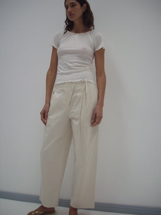 A woman with shoulder-length brown hair stands barefoot against a white wall, wearing a fitted white short-sleeve top and the Flag Tie Pant in Off White—relaxed, high-waisted, and made from organic cotton.