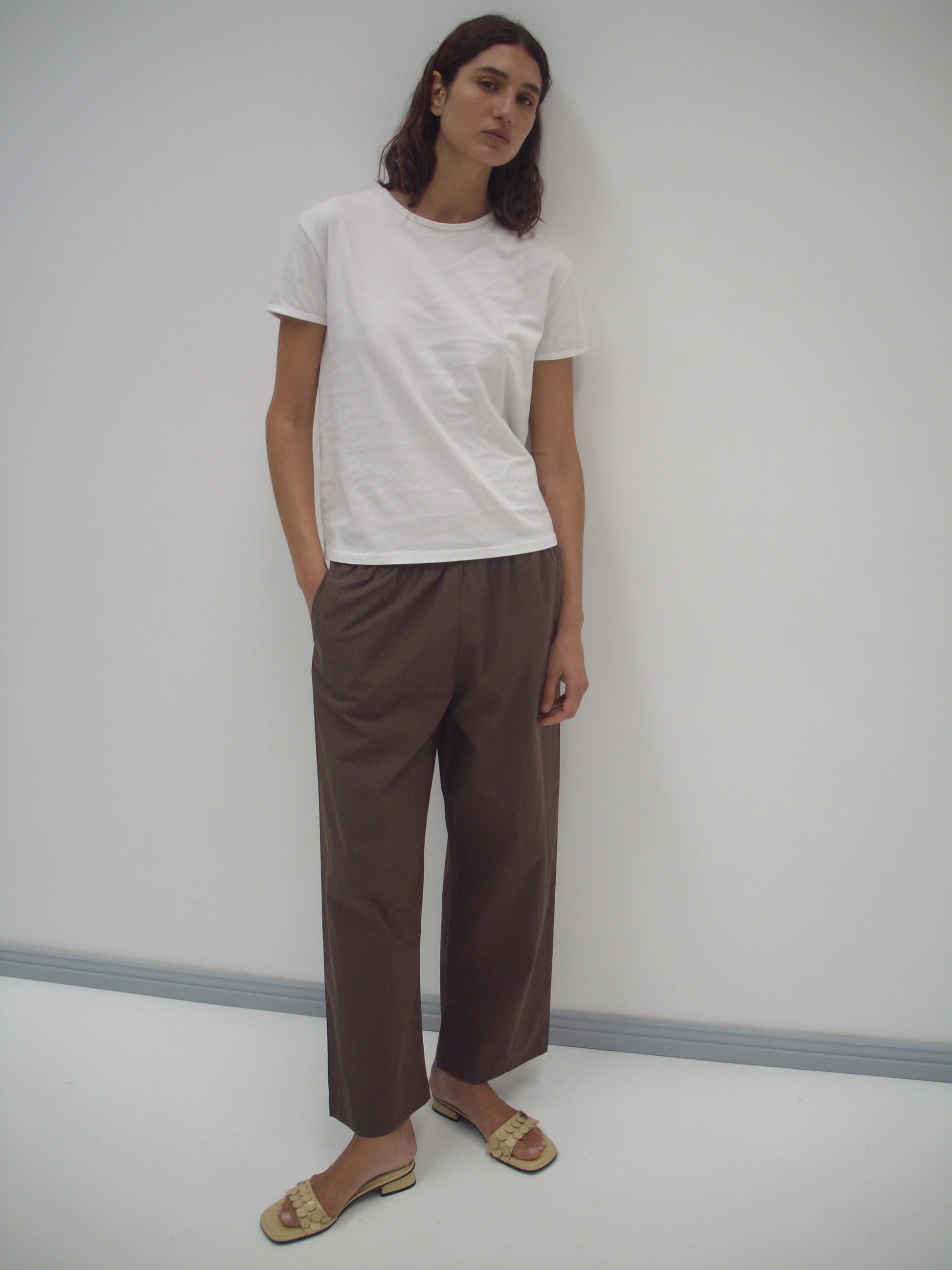 A person with long dark hair stands against a white wall, wearing the Simple Tee - White from Deiji Studios, loose brown trousers, and beige sandals. Their hands are in their pockets and they have a neutral expression.
