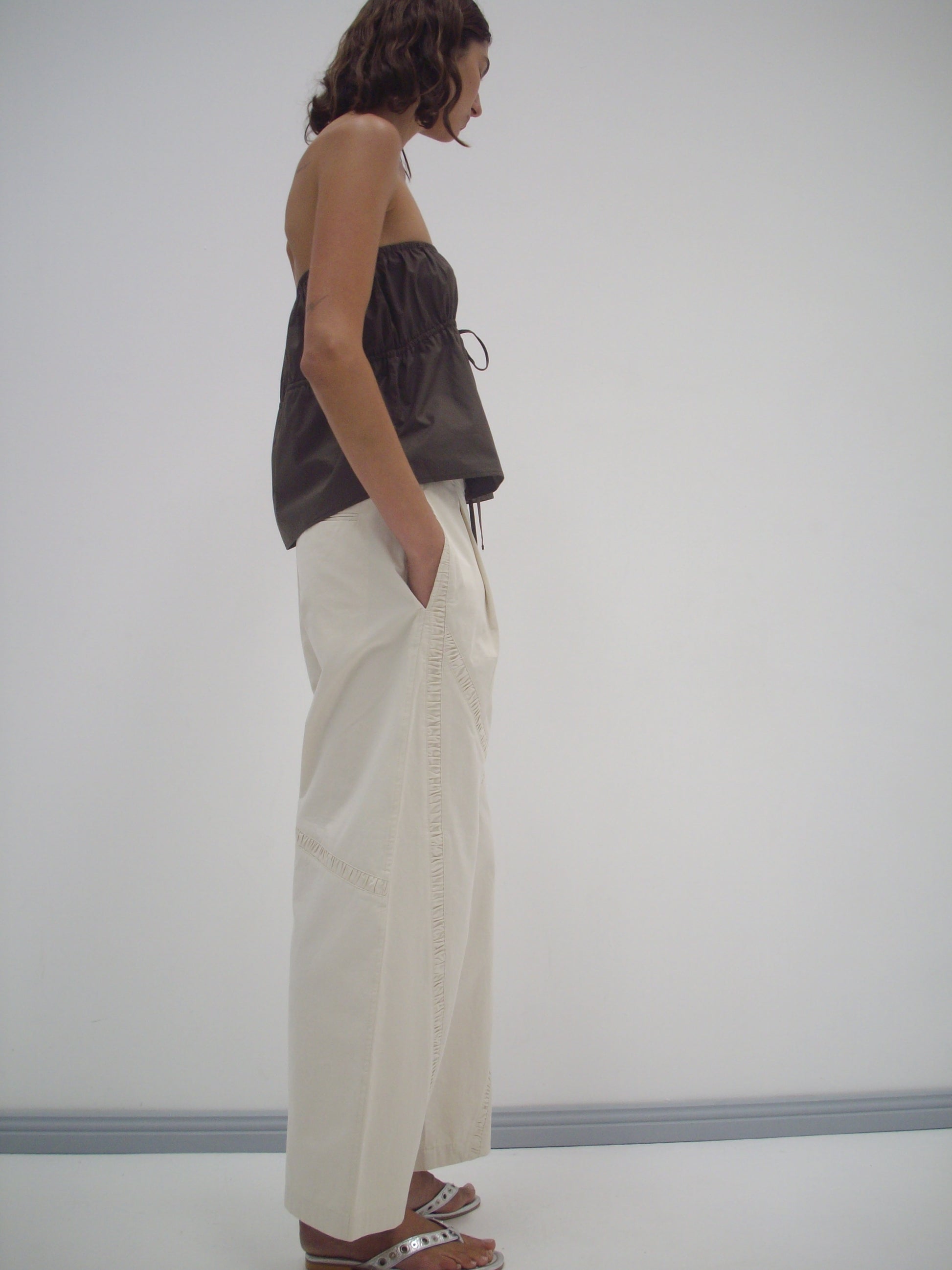 A woman stands in profile against a white wall, wearing Deiji Studios' Strapless Cotton Top in Chocolate, loose organic cotton white pants with pockets, and white sandals. Her hands are in her pockets and her wavy hair is shoulder-length.