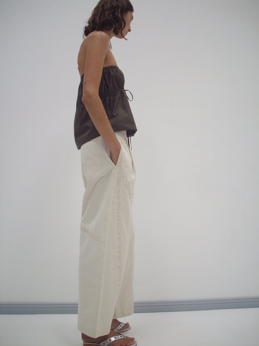 A woman stands in profile against a white wall, wearing Deiji Studios' Strapless Cotton Top in Chocolate, loose organic cotton white pants with pockets, and white sandals. Her hands are in her pockets and her wavy hair is shoulder-length.