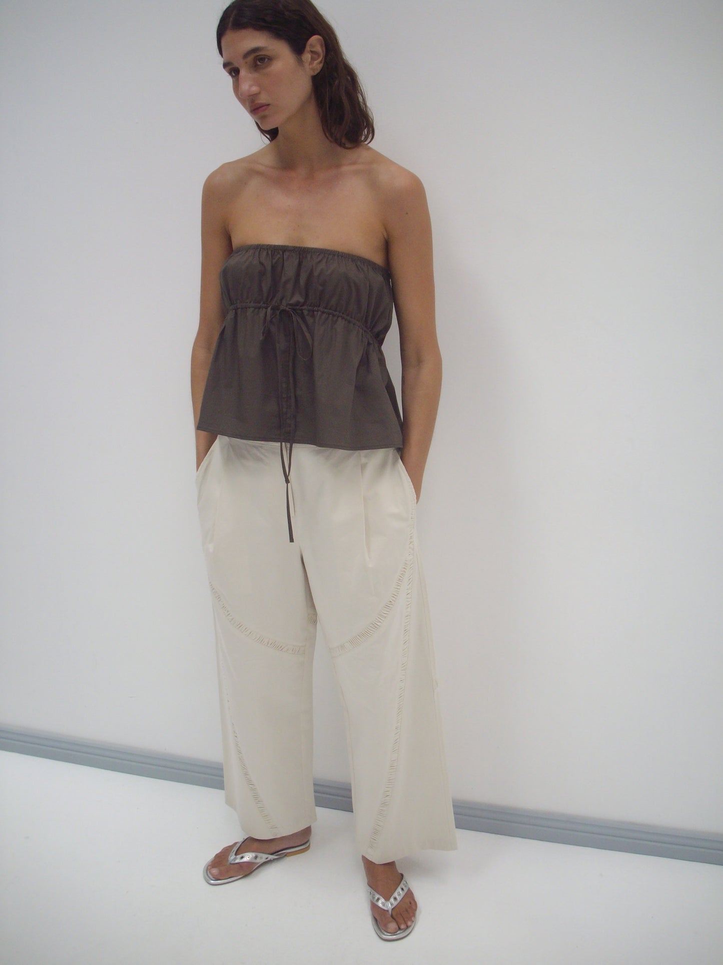A person stands in a white room wearing a Deiji Studios Strapless Cotton Top in Chocolate with loose, cream-colored organic cotton wide-leg pants and silver flip-flops. Their hands are in their pockets as they look off to the side.