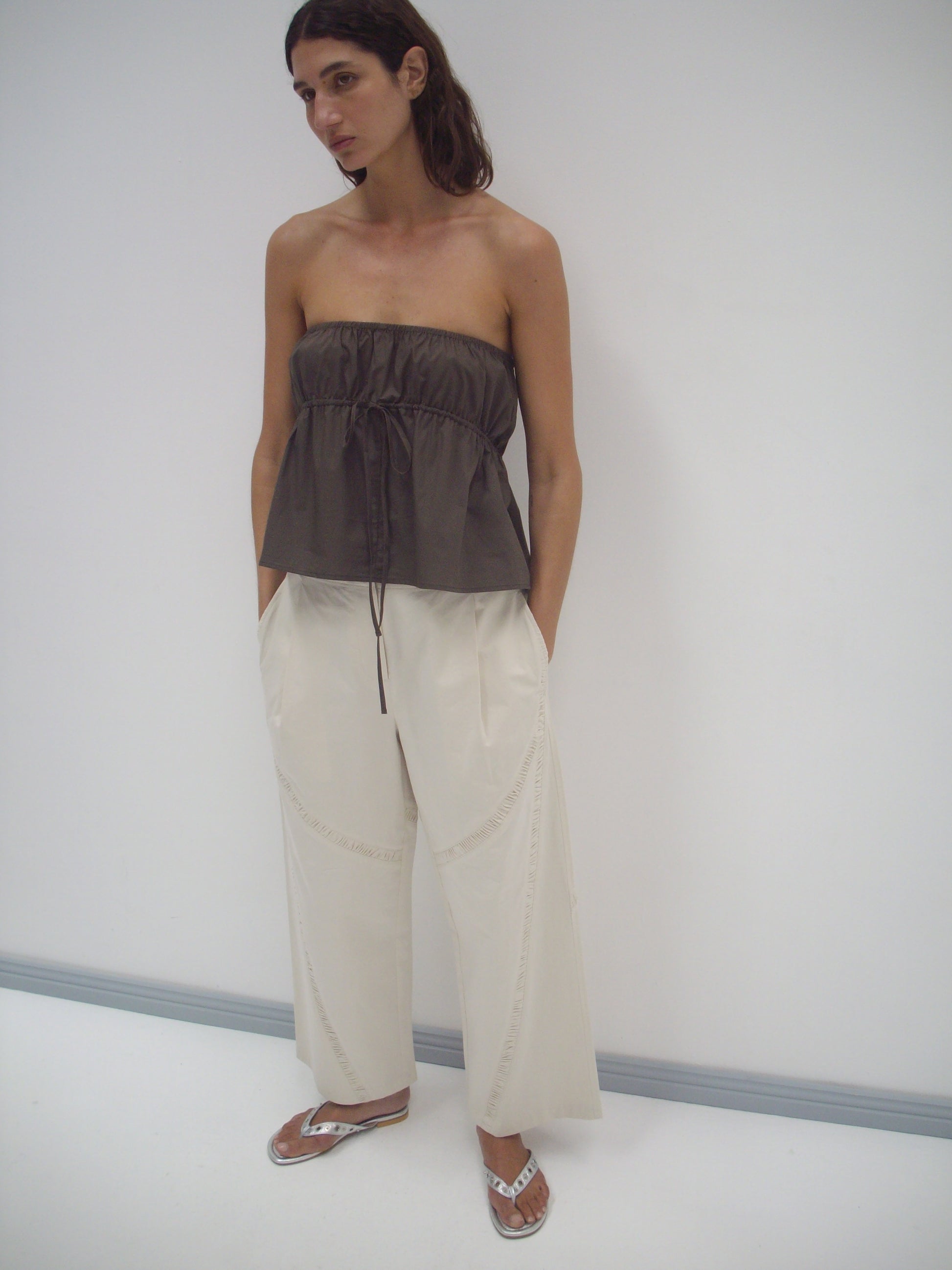 A person stands in a white room wearing a Deiji Studios Strapless Cotton Top in Chocolate with loose, cream-colored organic cotton wide-leg pants and silver flip-flops. Their hands are in their pockets as they look off to the side.