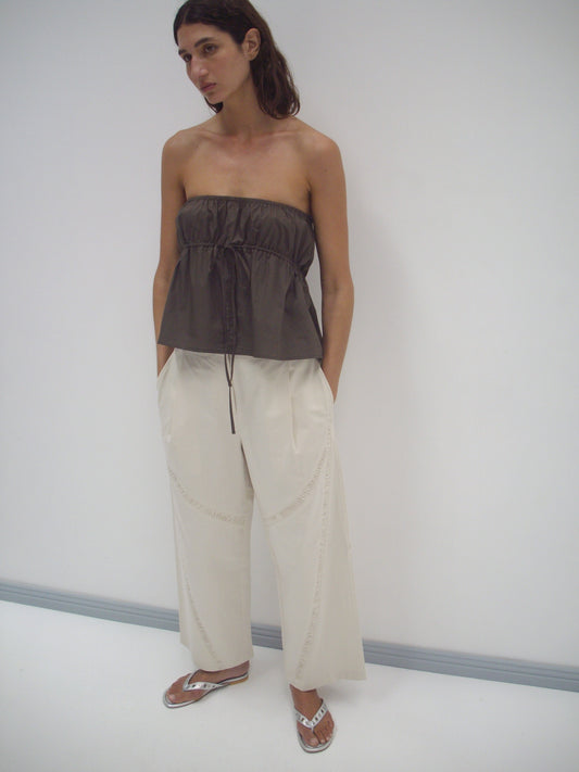 A person stands in a white room wearing a Deiji Studios Strapless Cotton Top in Chocolate with loose, cream-colored organic cotton wide-leg pants and silver flip-flops. Their hands are in their pockets as they look off to the side.