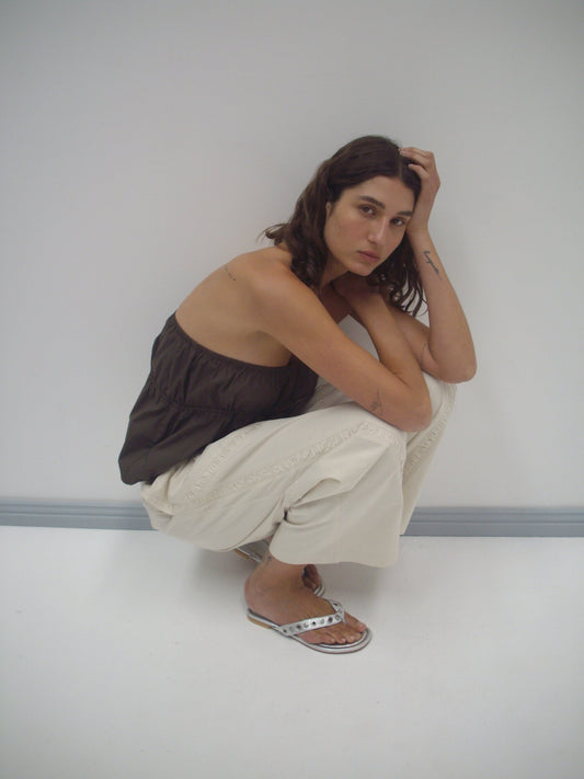A woman with wavy brown hair squats by a white wall, wearing the Strapless Cotton Top - Chocolate by Deiji Studios, light wide-leg organic cotton pants, and silver sandals, her elbow on her knee and hand resting on her head.