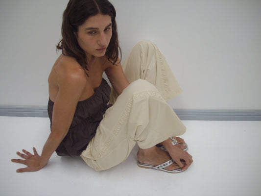 A person with long brown hair sits on the floor against a white wall, wearing a strapless brown top, Deiji Studios’ Channel Pant in Stone, and white sandals, looking off to the side with a neutral expression.