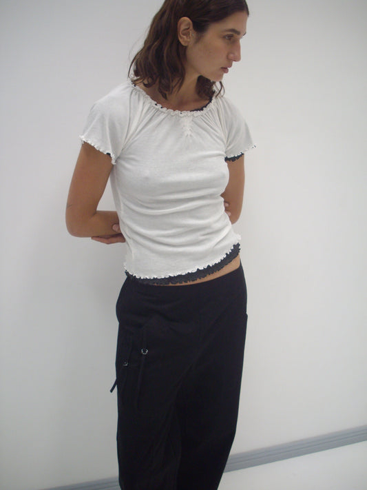 A person with medium-length brown hair stands against a white background, wearing a fitted white short-sleeve top and Deiji Studios Adjust Pocket Pant - Black in organic cotton, hands behind their back, looking to the side.