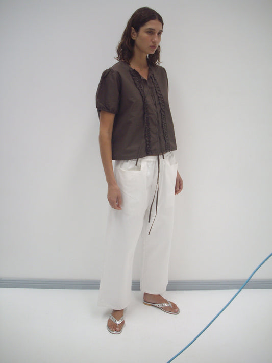A person with long brown hair wears a short-sleeve brown blouse, Square Pocket Pant - White in a wide-leg cut, and silver sandals, standing in a minimalist white room with a blue cord on the floor.