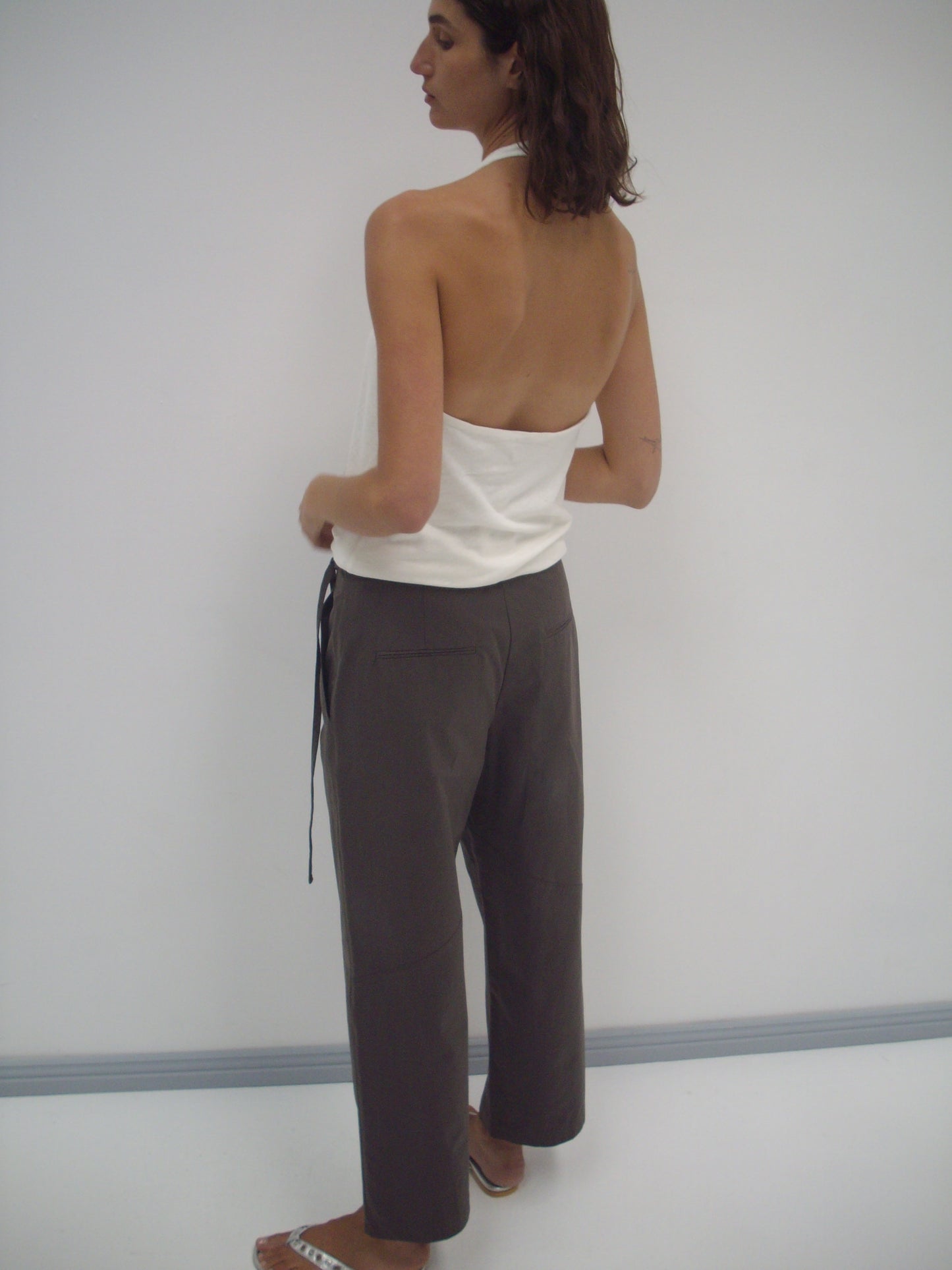 A person stands facing away, wearing the Flag Tie Pant in Dark Brown with a white halter top and white sandals, against a plain light-colored wall.
