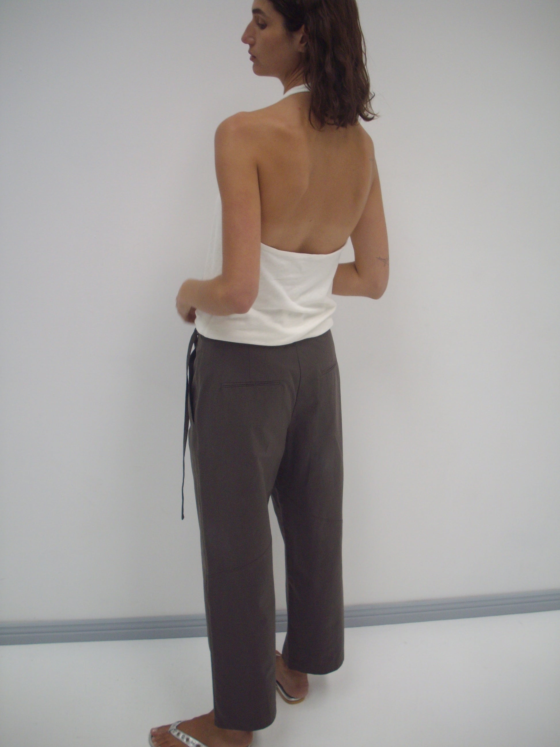 A person stands facing away, wearing the Flag Tie Pant in Dark Brown with a white halter top and white sandals, against a plain light-colored wall.