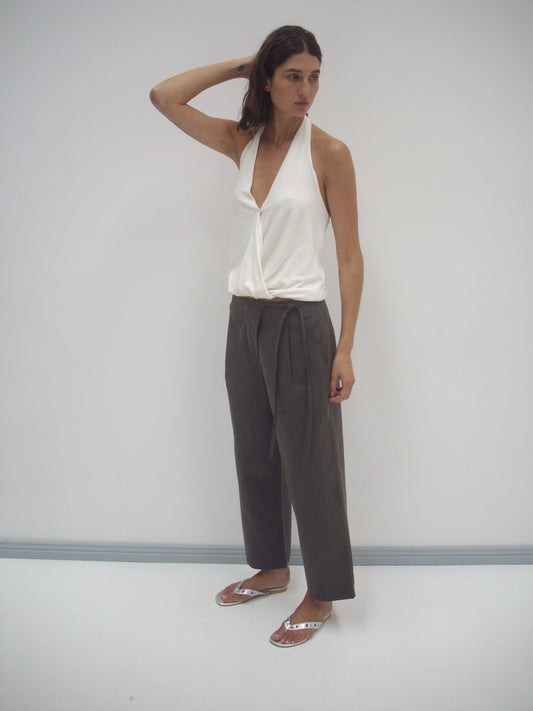 A woman stands against a plain white wall wearing a sleeveless, deep V-neck white top, Flag Tie Pant in dark brown, and silver sandals, with one hand raised to her head.