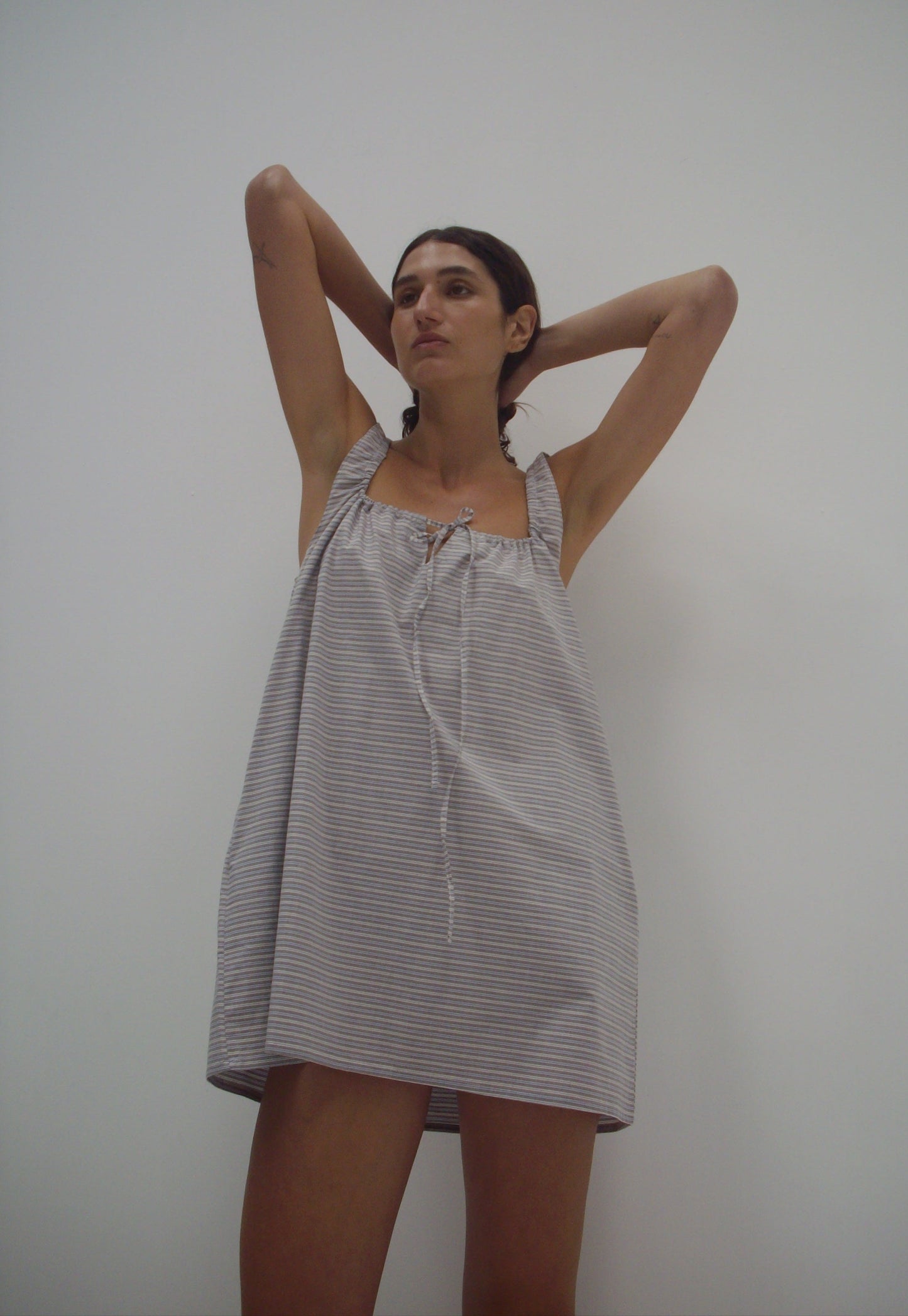 Wearing The Bow Dress - Horizontal Stripe, a loose, sleeveless mini dress made from organic cotton with gray stripes, a person stands against a plain white wall with raised arms and gazes slightly to the side.