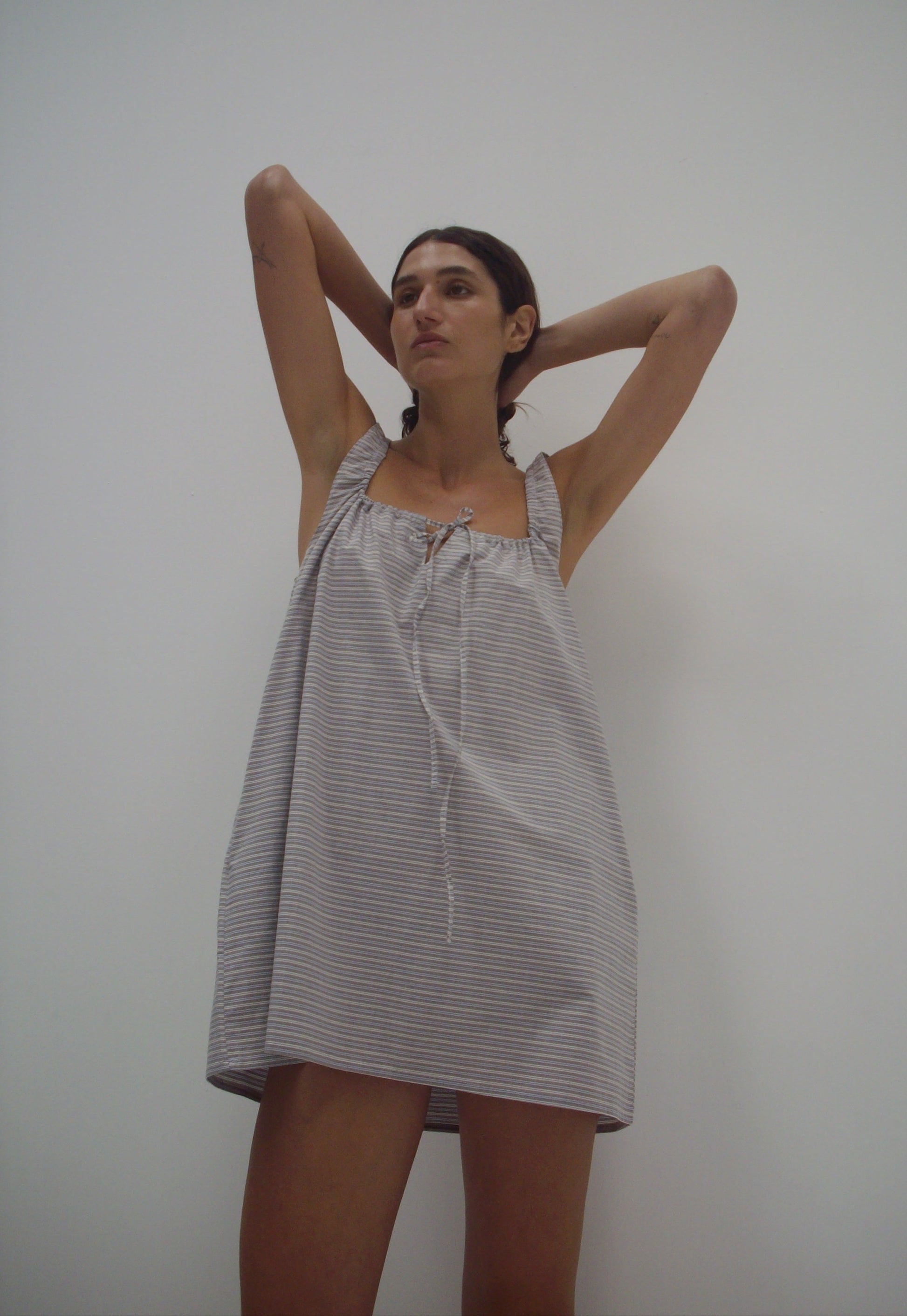 Wearing The Bow Dress - Horizontal Stripe, a loose, sleeveless mini dress made from organic cotton with gray stripes, a person stands against a plain white wall with raised arms and gazes slightly to the side.